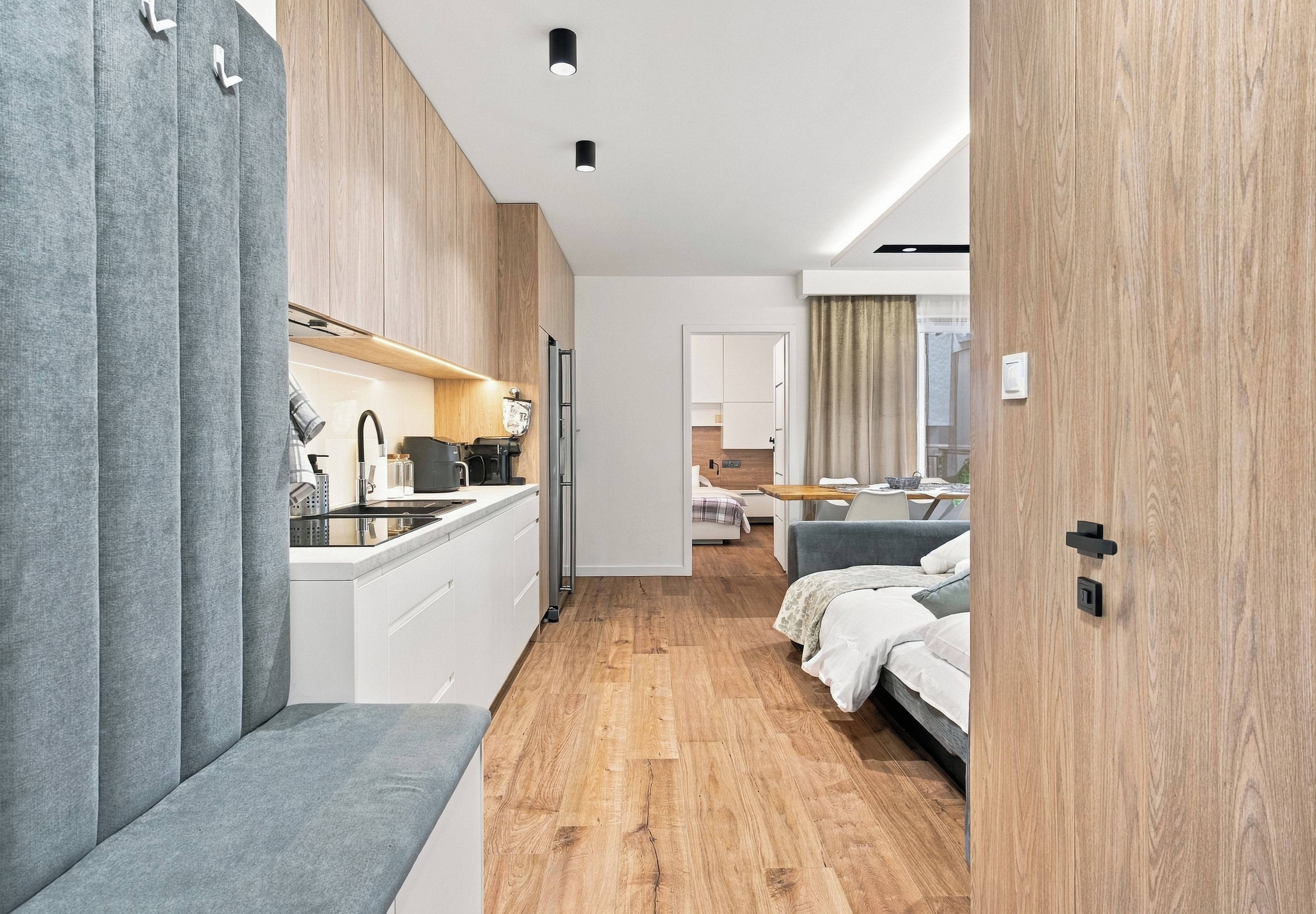 Une chambre d’hôtel moderne dotée d’une kitchenette, d’un plancher en bois, d’un lit et d’une salle de bain visible à travers une porte ouverte.