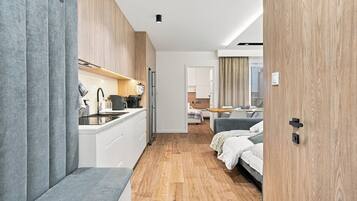 Une chambre d’hôtel moderne dotée d’une kitchenette, d’un plancher en bois, d’un lit et d’une salle de bain visible à travers une porte ouverte.