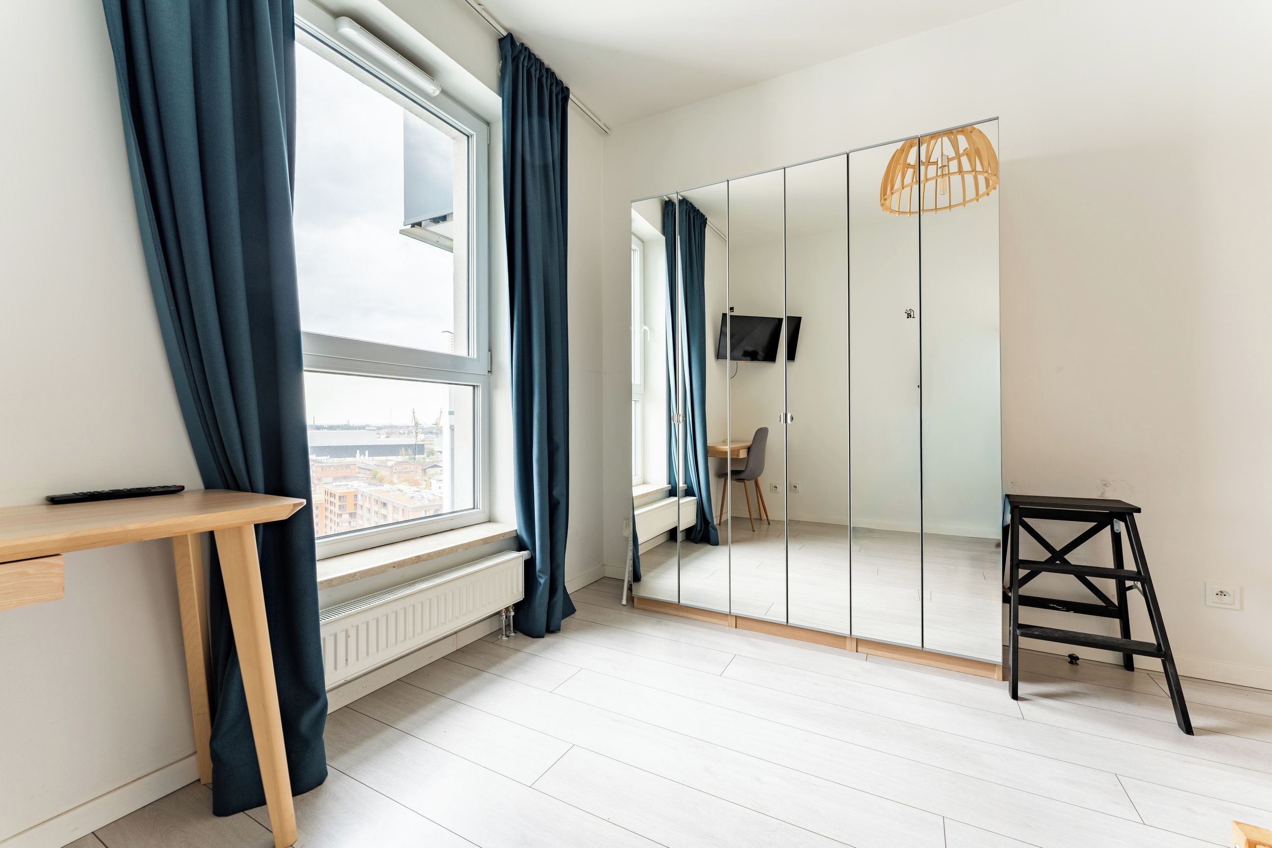 Une pièce avec une grande fenêtre, un bureau, une chaise, une armoire avec un miroir et un petit tabouret.