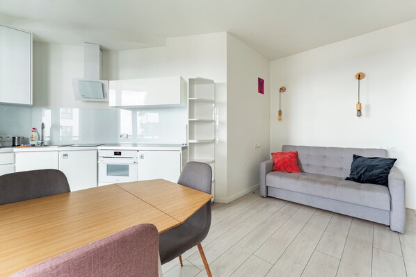 Eine moderne Küche mit weißen Schränken, einem eingebauten Backofen und einer Mikrowelle. Es gibt einen Esstisch mit Stühlen und ein graues Sofa mit roten und schwarzen Kissen.