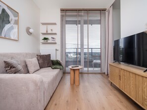 Ett modernt vardagsrum med en soffa, ett litet bord, en tv på ett träställ och en skjutdörr i glas som leder ut till en balkong.