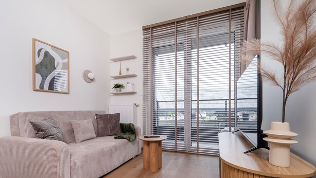 A modern living room with a sofa, a small side table, a decorative vase, and a large window with blinds. - Łódź Fabryczna Signature Apartment (Łódź)
