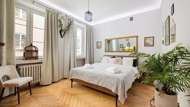 Ein Hotelzimmer mit Holzdielenmuster, einem Bett mit weißen Leinen, einem großen Spiegel und einer Topfpflanze.