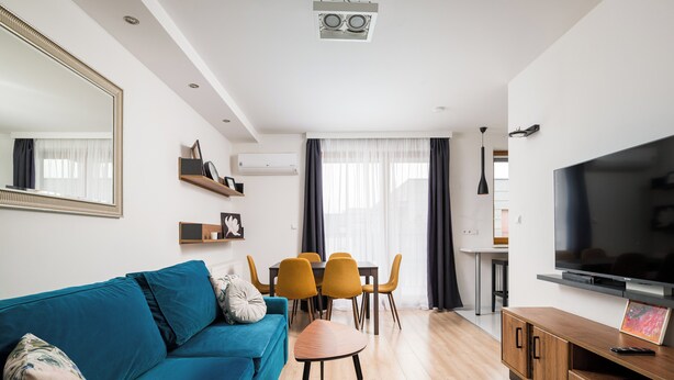 Ein modernes Wohnzimmer mit einem blauen Sofa, einem Esstisch mit Stühlen, einem Flachbildfernseher und einem großen Spiegel.