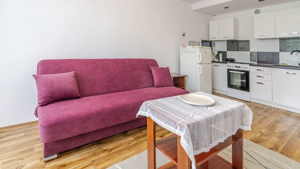 A modern living room with a pink sofa, a small table with a white lace tablecloth, and a kitchenette with white cabinets and a stove. - Piastów Standard Studio (Katowice)
