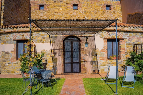 A stone building with a covered patio, outdoor seating, and a central entrance. - C/da Pozzetti Apartment (Lascari)