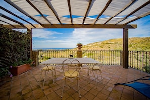 A covered patio with a table and chairs, a view of the countryside, and a stone pillar. - C/da Pozzetti Apartment (Lascari)
