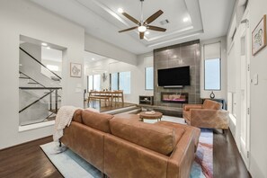 A modern living room with a brown leather sofa, a TV mounted on a stone wall, and a staircase leading to an upper level. - Luxury Retreat Near Memorial & Heights. Sleeps 8 with Wellness Studio (Houston)