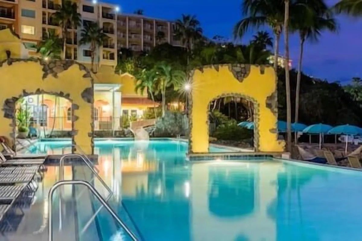 A swimming pool with clear water, surrounded by palm trees and a building with multiple balconies.