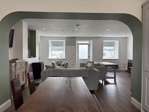 Een ruime woonkamer met een houten eettafel, een bank en een aan de muur bevestigde televisie.