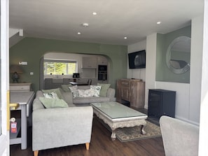 A living room with a sofa, coffee table, and fireplace. There is a kitchen in the background with cabinets and a TV mounted on the wall. - Cottage 519 - Kilkieran - sleeps 6 guests  in 3 bedrooms (Kilkerrin)