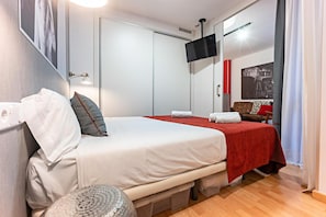 Et hotelværelse med en seng, et lille rundt bord, et fjernsyn ophængt på væggen og en dør med håndtag.