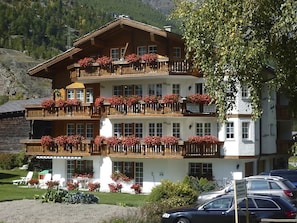 Ein mehrstöckiges Hotel mit Balkonen, die mit Blumen geschmückt sind, ein Parkplatz und eine gebirgige Kulisse.