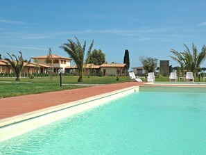 A swimming pool with clear water, surrounded by a brick walkway. There are lounge chairs and palm trees in the background. - A5 - Three Bedroom (Grosseto)