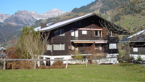 Un chalet à deux étages doté d’un balcon, entouré d’une cour verdoyante et avec des montagnes en toile de fond.