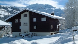 Un chalet à deux étages, doté d’un toit en pente, entouré de montagnes et d’arbres couverts de neige.