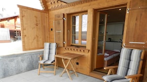Une cabane en bois avec deux chaises et une table, une porte donnant sur une chambre à coucher, et une vue sur un bâtiment à l’extérieur.