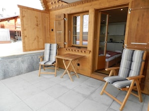 A wooden cabin with two chairs and a table, a door leading to a bedroom, and a view of a building outside. - Mutthorn - One Bedroom (Lauenen)