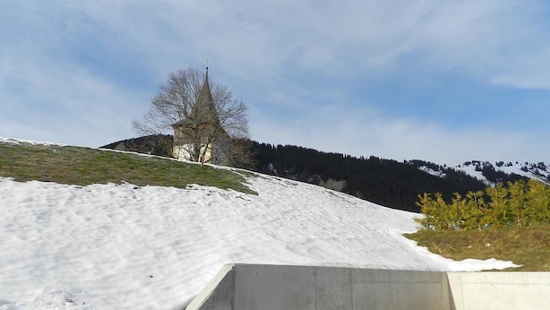 Un paysage enneigé avec un bâtiment perché sur une colline, un ciel dégagé et une structure en béton au premier plan.
