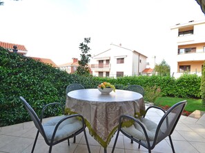 A round outdoor table with a fruit bowl, surrounded by chairs with cushions, set on a tiled patio with a view of residential buildings and greenery. - Stana - One Bedroom (Zadar)