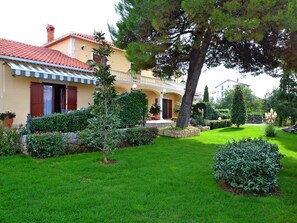 A two-story house with a red-tiled roof, a balcony with a railing, and a well-maintained garden with various shrubs and a lawn. - Stana - One Bedroom (Zadar)