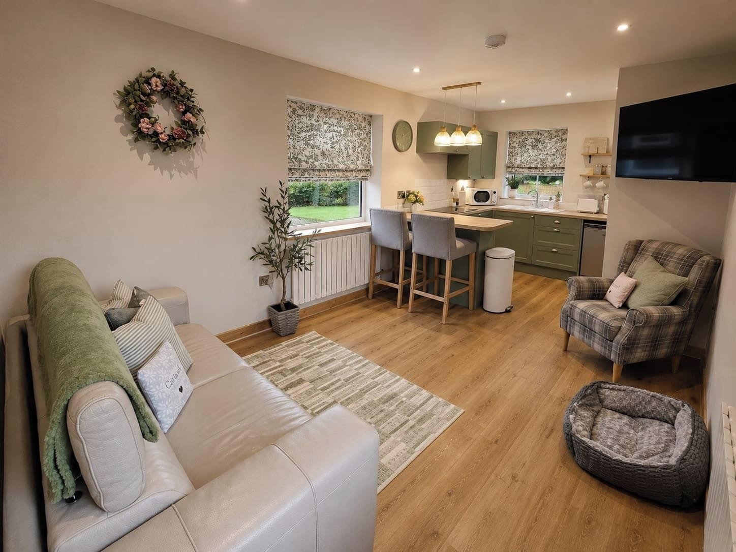 A modern living room with a sofa, a dining area with chairs, a kitchen with a microwave, and a TV mounted on the wall.