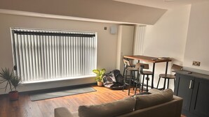 Una sala moderna con sofá, mesa de comedor con sillas, planta en maceta y ventana con persianas.