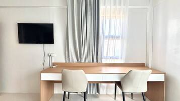 A modern room with a long wooden desk, two chairs, a flat-screen TV mounted on the wall, and a window with sheer curtains.