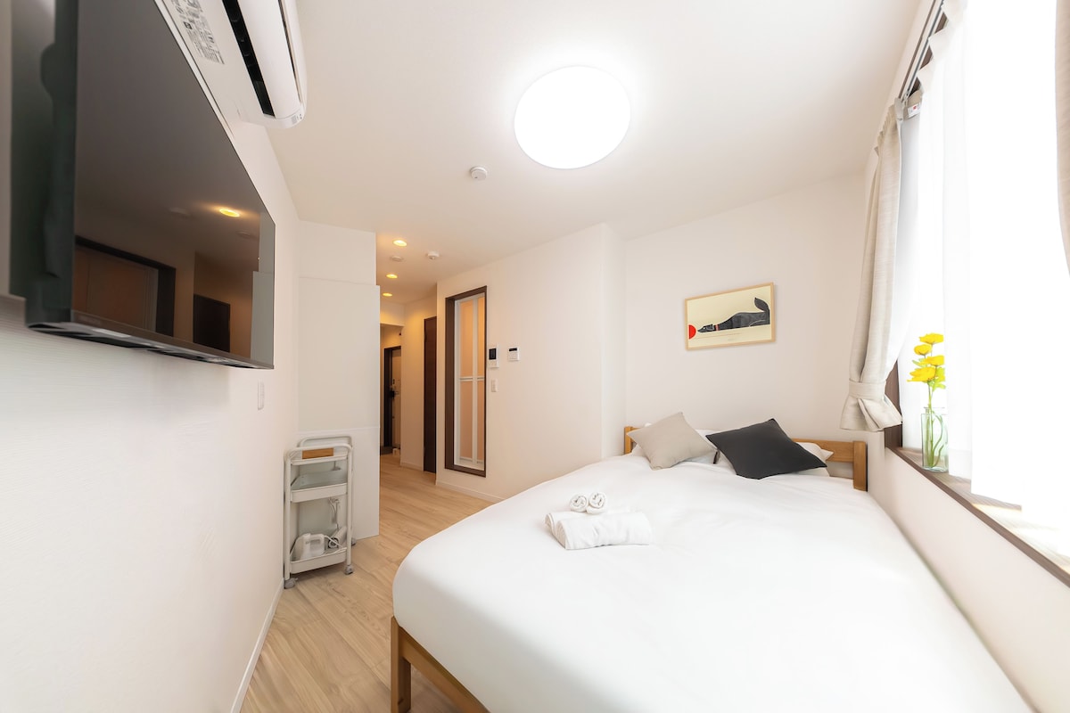 Ein Hotelzimmer mit Bett, Nachttisch, einem Fenster mit Vorhängen, einer Wandklimaanlage, einer Tür zu einem anderen Raum und einem kleinen Nachttisch mit Blumen.