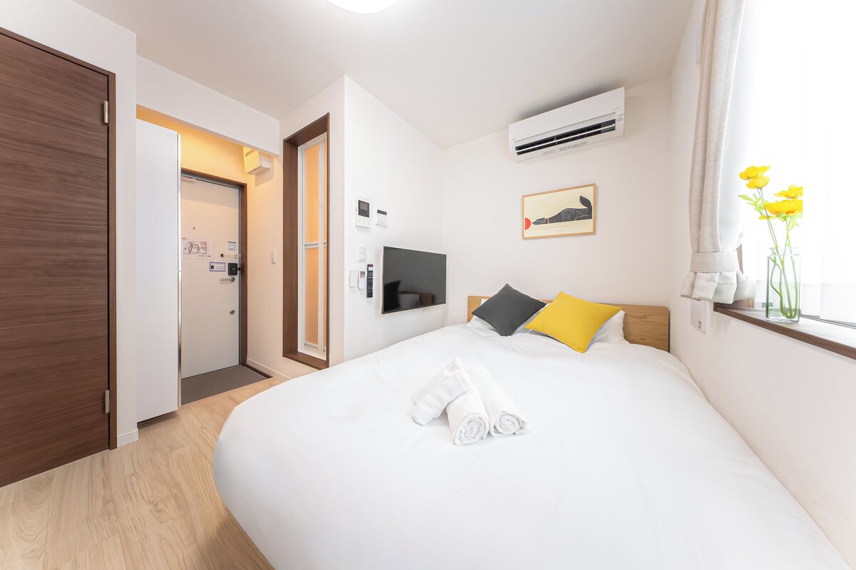 Ein Hotelzimmer mit Bett, Fernseher, Klimaanlage, einem Gemälde und einem Fenster mit Blumen.