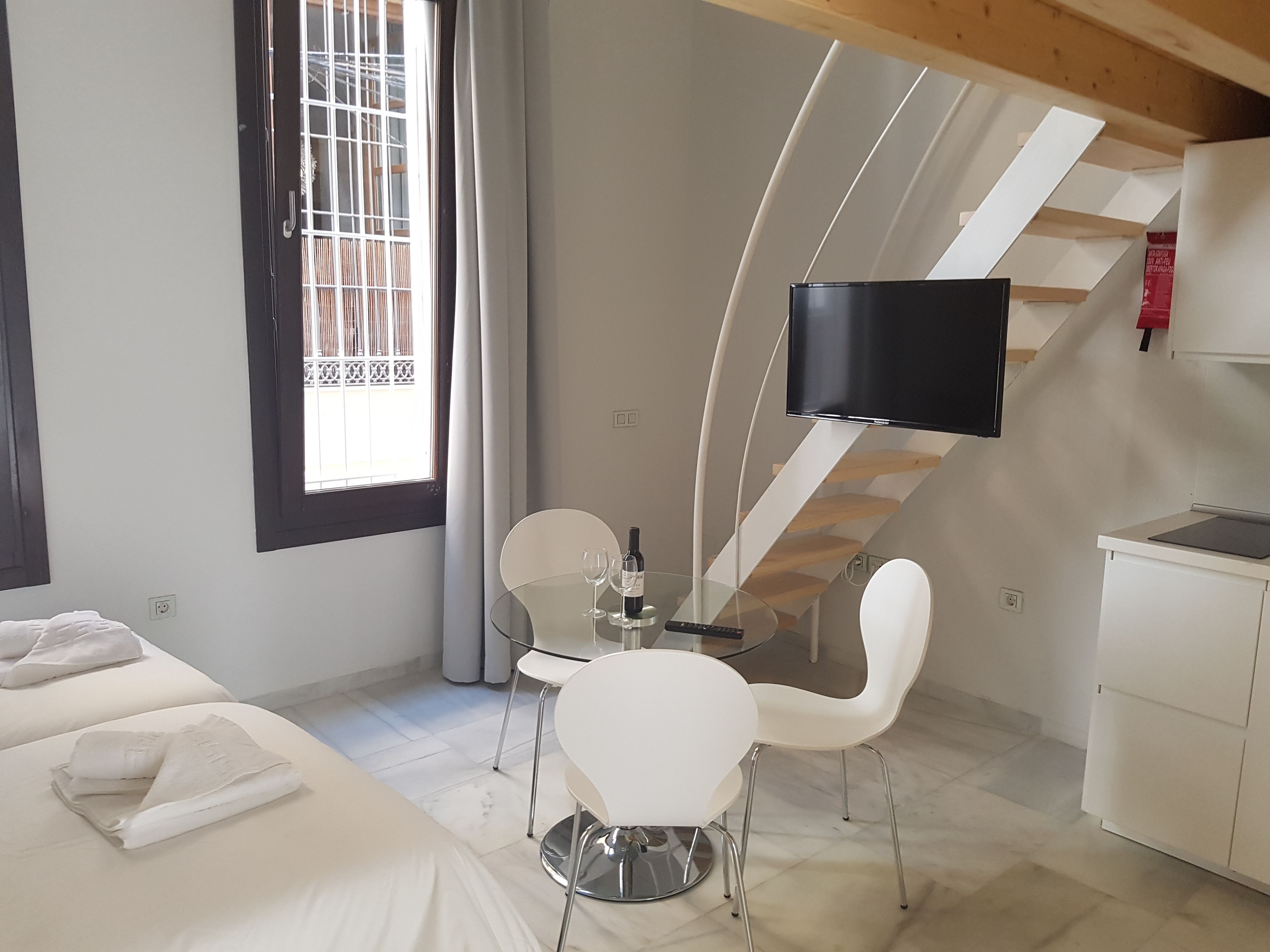 A modern hotel room with a bed, a dining table with chairs, a TV mounted on a wall, and a staircase.
