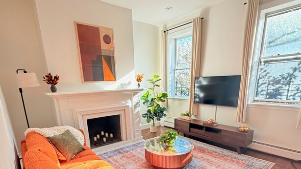 A living room with a sofa, a fireplace, a TV, and a large window. - Charming Historic 2BR Townhouse 10-Min PATH Ride to NYC (Jersey City)