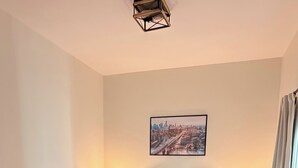 A bedroom with a bed, two bedside tables, a cityscape painting, and a window with curtains. - Charming Historic 2BR Townhouse 10-Min PATH Ride to NYC (Jersey City)