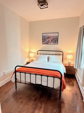 Une chambre à coucher avec un lit, deux tables de chevet, un tableau représentant un paysage urbain et une fenêtre avec des rideaux.