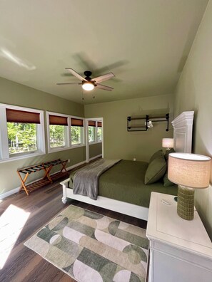 Un dormitorio con cama, ventilador de techo, mesita de noche con lámpara, ventana con persianas y banco de madera.