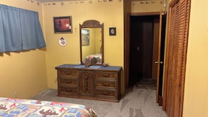 A bedroom with a wooden dresser, a bed with a colorful bedspread, a mirror, and a door leading to another room.