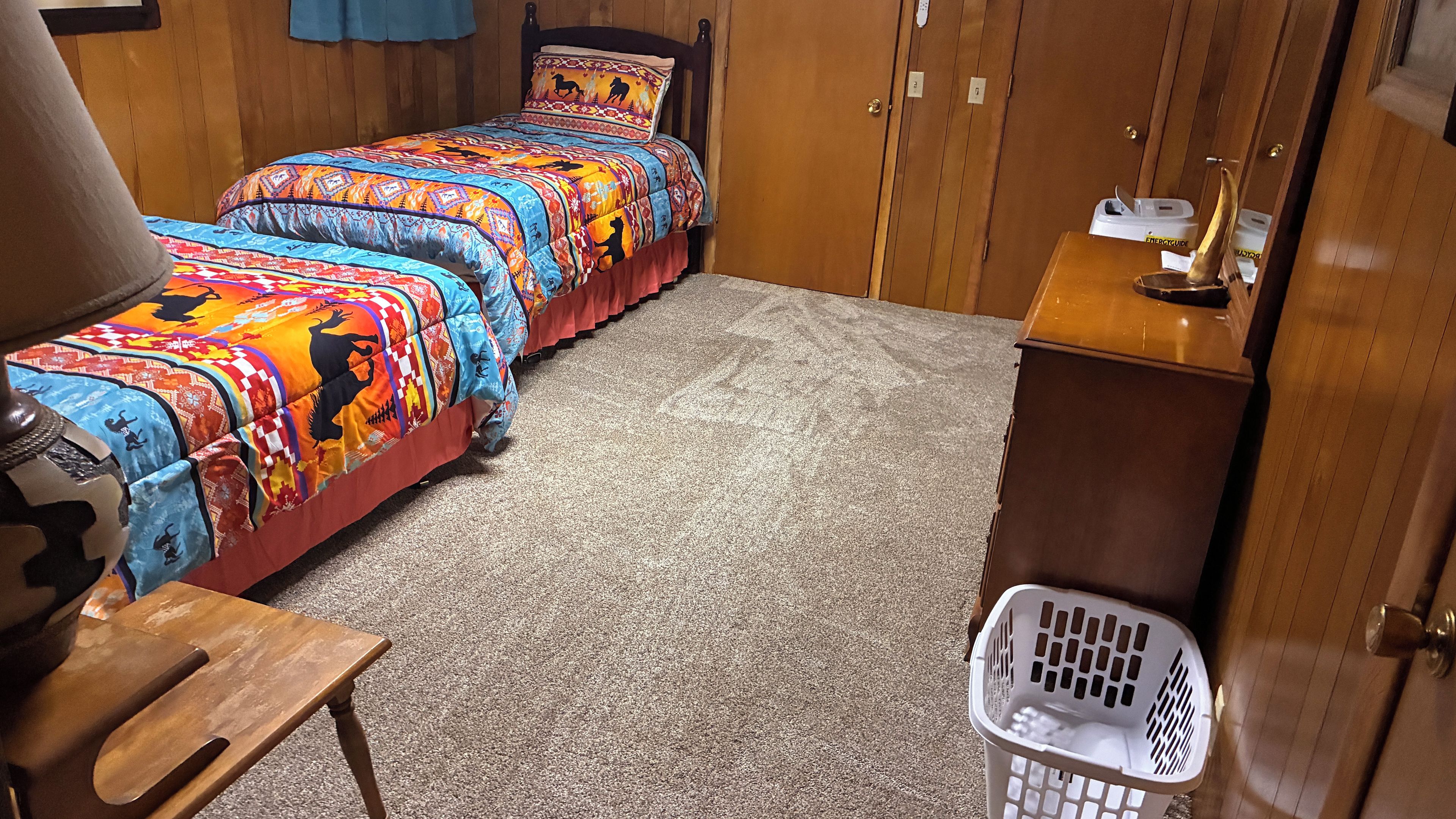 A bedroom with two beds, a wooden headboard, a small wooden table, a laundry basket, and a wooden cabinet.