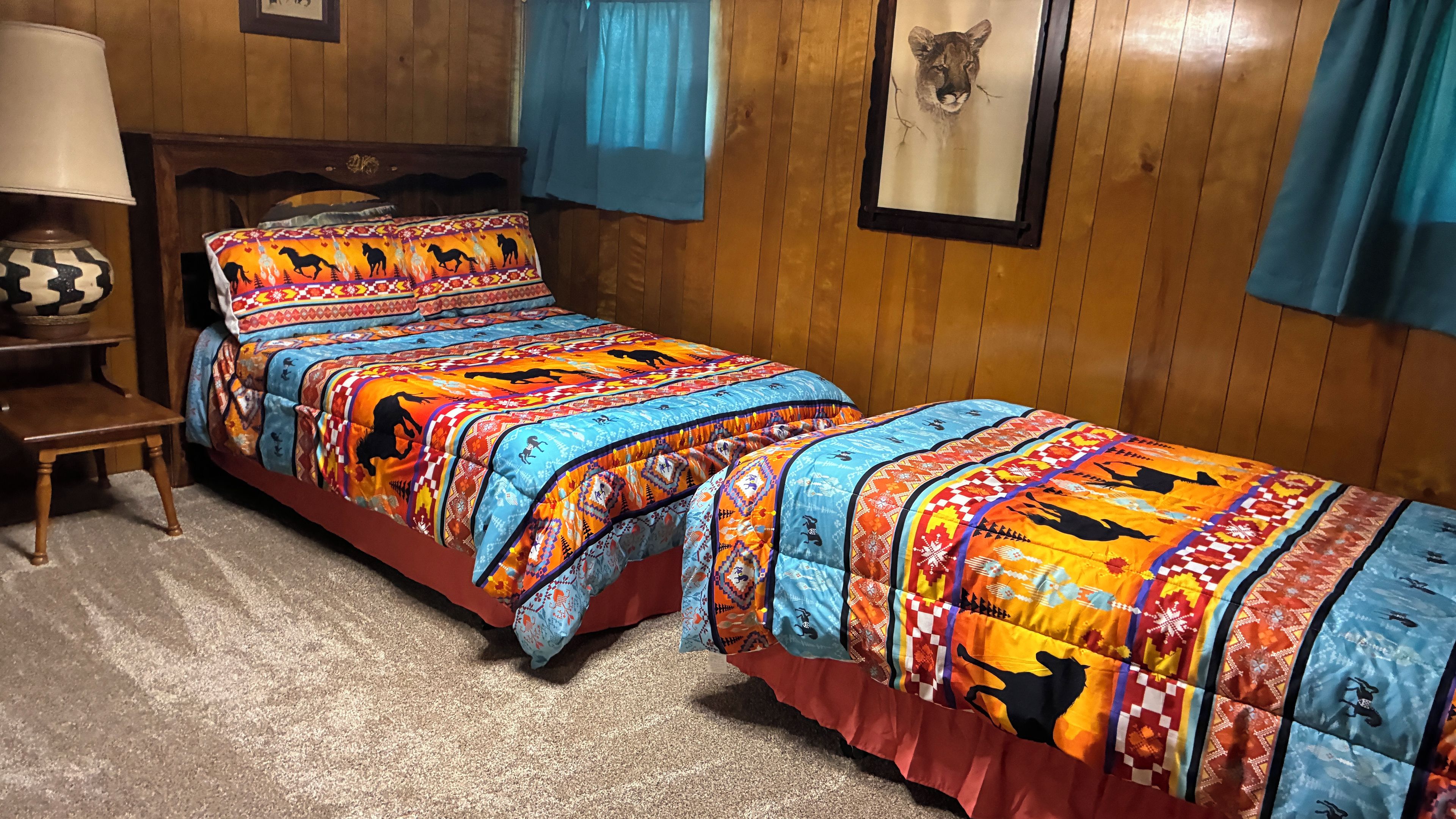 A room with two beds, a wooden headboard, a nightstand, a lamp, and a framed picture on the wall.