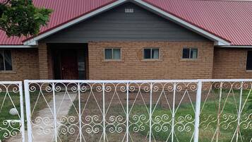 Uma casa de dois andares com telhado vermelho, paredes de tijolo e cerca de metal ao primeiro plano.