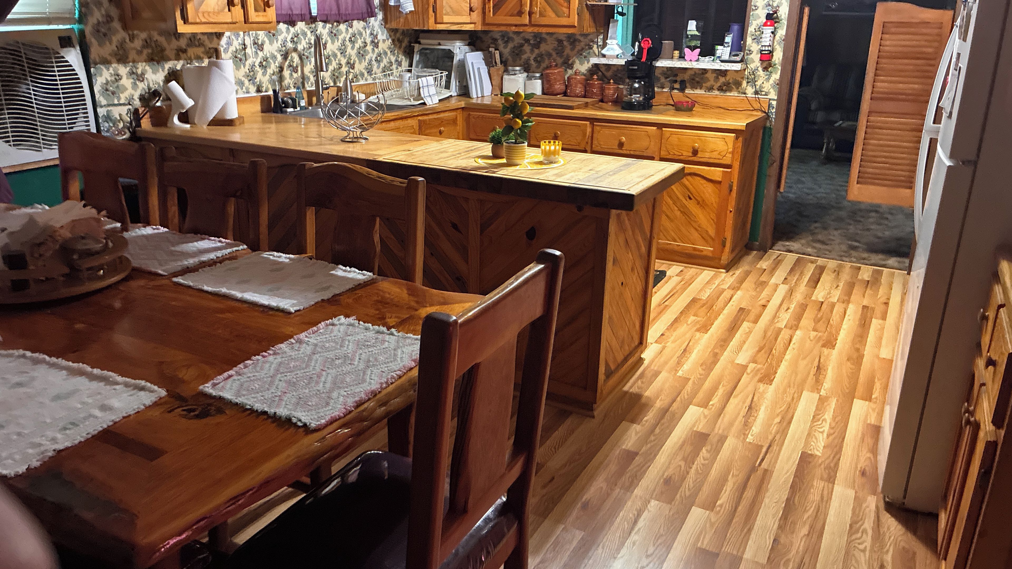 A wooden dining table with placemats, a kitchen area with wooden cabinets, and a wooden floor.