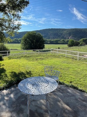 Un tavolo rotondo in metallo con decorazioni elaborate, una sedia in metallo e vista su un prato verde con alberi e colline sullo sfondo.