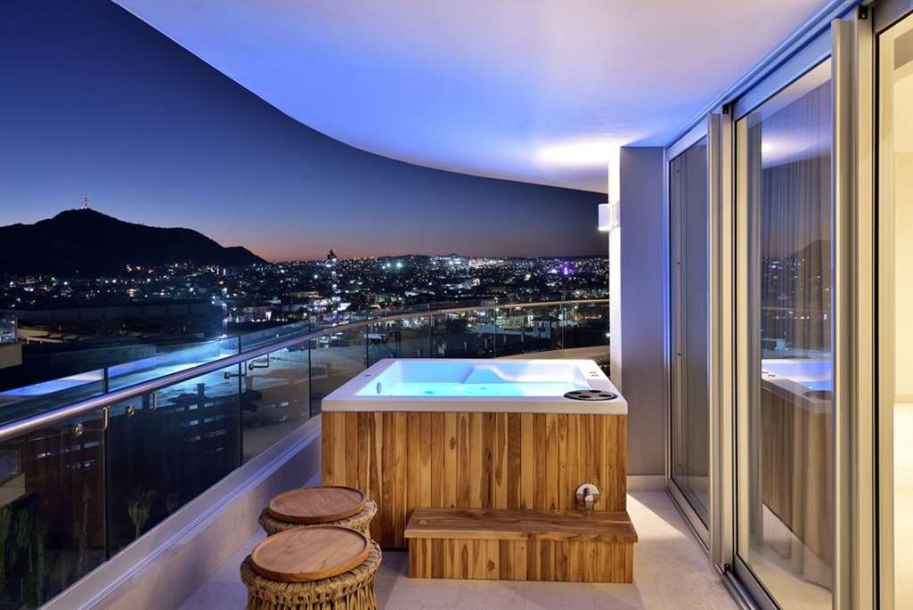 A balcony with a hot tub, wooden stools, and a city view at dusk.