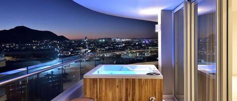 A balcony with a hot tub, wooden stools, and a city view at dusk.