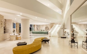 Eine großzügige Lobby mit einer zentralen Rezeption, einer gebogenen gelben Couch und einer Treppe zu den oberen Stockwerken.