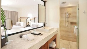 Une salle de bain d’hôtel moderne avec un meuble-lavabo à deux vasques, un grand miroir, un espace douche et un lit avec une tête de lit.