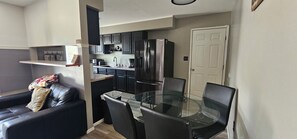 A compact kitchen with dark cabinets, a stainless steel refrigerator, and a dining area with a glass table and chairs.