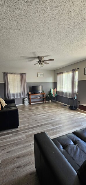 A spacious living room with a ceiling fan, a flat-screen TV, a black leather sofa, and a wooden coffee table.