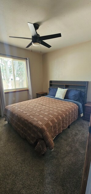 Ein Schlafzimmer mit einem Bett, einem Deckenventilator, einem Nachttisch, einem Fenster mit Jalousien und einem Teppichboden.