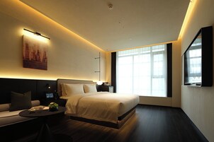 Una habitación de hotel moderna con una cama grande, una zona de estar con una mesa y un ventanal que proporciona luz natural.