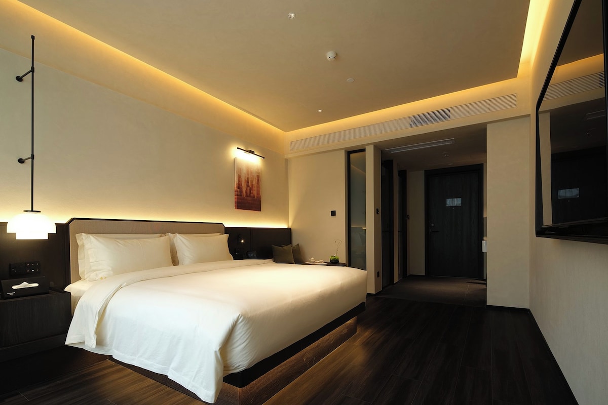 A modern hotel room with a large bed, bedside tables, a wall-mounted TV, and a hallway leading to another room.